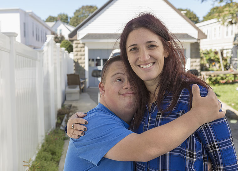 Smiling young man with disability hugging and looking up at smiling woman, standing outside in front of a garage