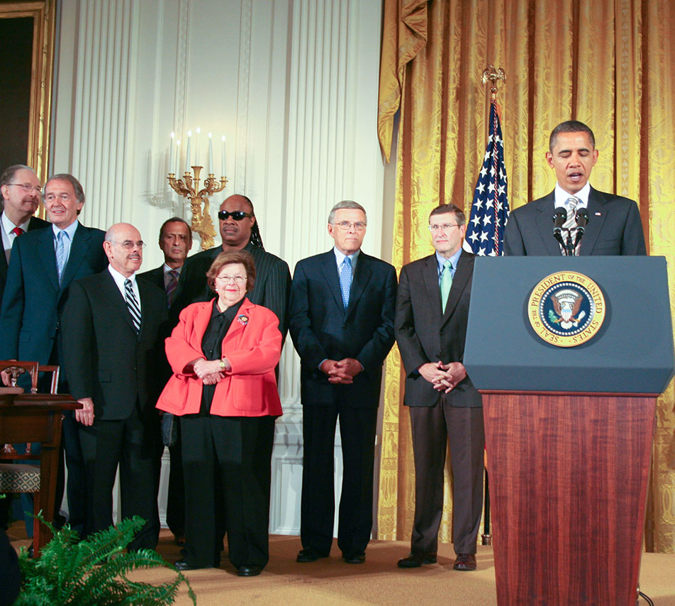 President Obama giving a speech while standing behind the presidential podium, with group of people standing behind him